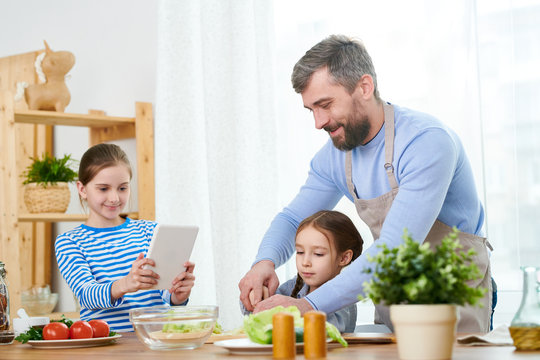 Smiling middle-aged man wearing apron teaching his little daughter to prepare vegetable salad while gathered together at cozy kitchen, her elder sister studying recipe on digital tablet