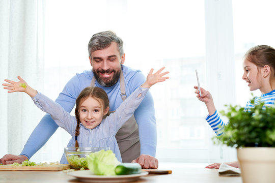 Profile view of smiling little girl taking picture of her handsome dad and cute sister while they wrapped up in preparing vegetable salad, kitchen interior on background