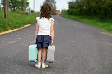 Une petite fille de dos, sur la route, avec une valise dans chaque main, les pieds serrés