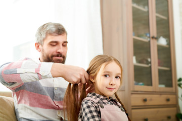 Bearded middle-aged man sitting on couch and braiding hair of his adorable little daughter, she looking at camera with warm smile
