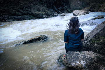 A girl sits with her back on the bank of a mountain river