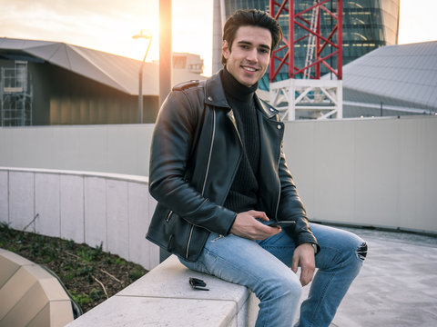 One handsome young man in urban setting in modern city, sitting, wearing black leather jacket and jeans