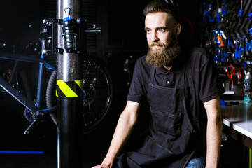 Portrait of small business owner of young man with beard. Guy bicycle mechanic workshop worker sitting with tool in his hand in a working black clothes in an apron in the background of a bicycle shop.