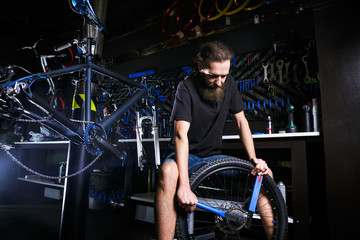 Fototapeta na wymiar Young stylish bearded, with long hair Caucasian male mechanic worker at a bicycle workshop uses a tool for repairing Cassettes on the rear wheel of a bicycle