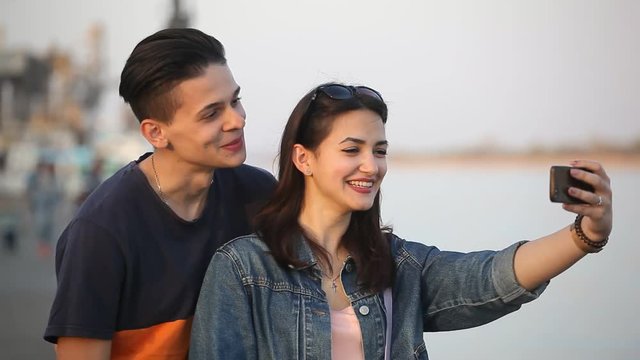 Portrait of a lucky young man kissing his lovely sweetheart girl on the Dnipro embankment in spring in slow motion. They take an amorous selfie 