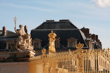 Golden royal crown, fence, white marble group sculpture and the roof of Versailles palace under blue sky