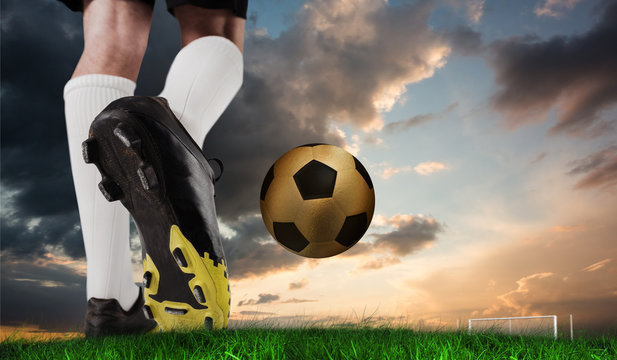 Composite image of football boot kicking gold ball against green grass under blue and orange sky