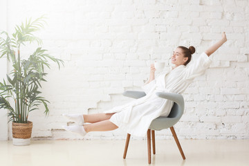 Studio photo of young good-looking European Caucasian female pictured in bright white room dressed in white bathrobe stretching having just woken and got up in morning getting ready for new day