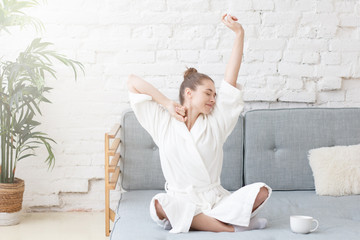 Horizontal photo of young beautiful Caucasian woman sitting on gray couch in bright white room stretching in white bathrobe in morning drinking coffee trying to wake up and start new good day - obrazy, fototapety, plakaty