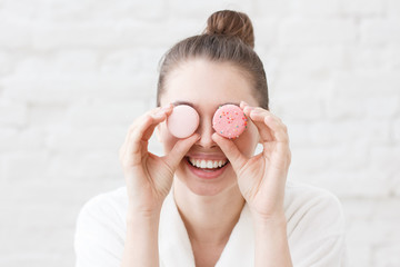 Indoor closeup of young good-looking European Caucasian female pictured in white bathrobe against white brick background holding colorful macarons in front of eyes having much fun feeling excited