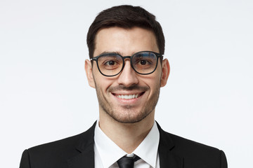 Closeup headshot of young handsome European businessman in glasses pictured isolated on gray background wearing black formal suit, white shirt and tie, looking confident and smiling friendly
