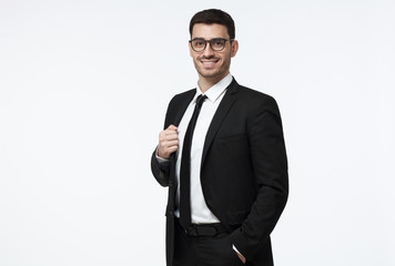 Half-length portrait of handsome Caucasian guy dressed in black suit, white shirt and tie isolated on gray background looking attentively through glasses and smiling positively and confidently