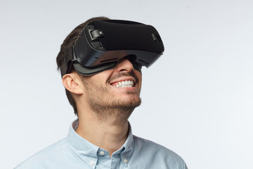 Horizontal closeup of young European Caucasian man with dark face hair isolated on grey background dressed casually, having VR headset on, experiencing its facilities with absolute satisfaction