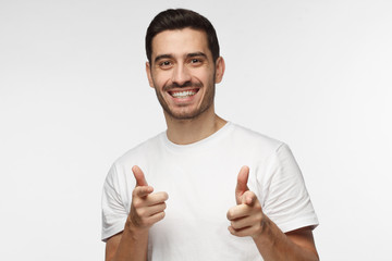 Closeup portrait of young happy European Caucasian man isolated on gray background smiling while looking at camera pointing to viewer with fingers of both hands as if approving and feeling pleased