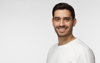 Closeup photo of young handsome European man pictured isolated on grey background dressed in white casual T-shirt in right side of picture smiling, copyspace for advertisement on left side of photo