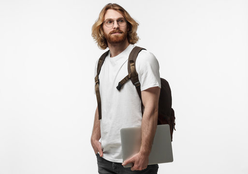 Half-length horizontal picture of young hipster long-haired man wearing glasses isolated on white background dressed in white T-shirt, wearing backpack, holding closed laptop, ready to go for walk