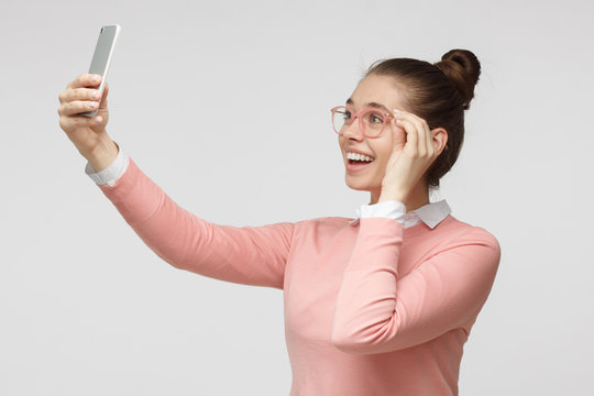 Horizontal photo of young European female in casual clothes and glasses isolated on grey background taking selfie picture with cellphone smiling happily as if expecting to get likes in social networks