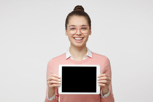Indoor photo of young beautiful European female isolated on gray background showing blank screen of tablet computer she is showing to advise something, with copyspace of products and services