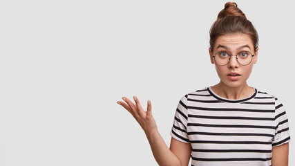 Indoor shot of amazed beautiful young female customer being shocked with prices, wears striped casual t shirt, wants to buy new outfit, gestures with hand as shows something at blank copy space