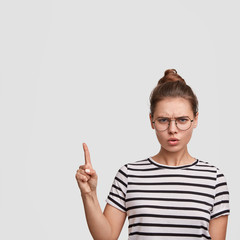 Angry Caucasian young female in striped t shirt and round glasses, indicates with displeased expression upwards, being discontent with something, poses against white backgroud, copy space for text