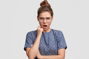 Portrait of annoyed shocked discontent beautiful female expresses irritation and displeasure, reacts on bad news, wears spectacles and polka dot blouse, poses against white studio background