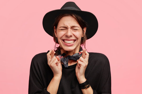 Portrait of stressful annoyed female clenches teeth and crosses fingers in anticipation, has desperate expression, wears stylish black hat, poses against pink background. Please let`s dreams come true