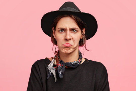 Horizontal shot of discontent annoyed young female model curves lips and has unhappy expression, wears black sweater and hat, poses against pink background. Irritated cowgirl shows her dislike