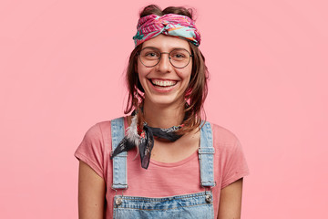 Glad positive female hippie happy to belong to this youth subculture, has specific appearance, wears indian style of clothes, has broad smile on face, isolated on pink background. Hippy woman