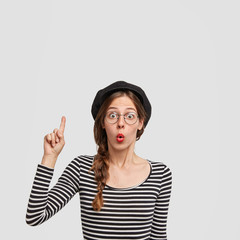 Vertical shot of surprised young female teacher of French in elegant beret and striped sweater indicates at blank copy space, shows something to pupils, blank copy space for your advertisement
