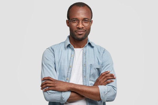 Image of confident attractive young African American male with pleased expression, keeps hands crossed, wears denim shirt and round spectacles, being content to recieve praisement from boss.