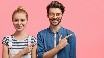 Positive European friends dressed in fashionable clothes, stand next to each other, point aside at blank copy space for your advertising content. Pretty blonde woman with hair bun and hipster guy