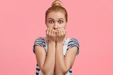 Beautiful nervous blue eyed female with light hair tied in knot, bites finger nails, feels anxious as going to make speech in front of audience, isolated over pink background. Puzzled worried girl