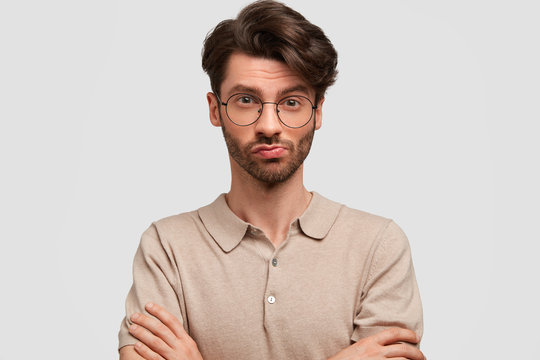 Serious confident bearded male with pleasant appearance, keeps arms folded, looks with mysterious expression at camera, wears big spectacles, isolated on white background. Facial expressions concept