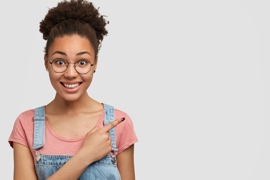 Horizontal shot of good looking cheerful mixed race female has positive expression, curly dark hair, wears casual clothes, indicates at blank copy space for your promotional text or hearder.