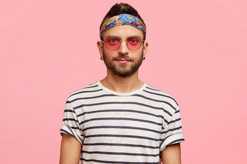 Horizontal portrait of serious bearded young male hippie in stylish sunglasses, headband and...