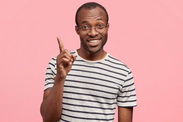 Delighted dark skinned male with positive smile, has good idea in mind, raises fore finger, wears casual striped t shirt, isolated over pink background. People, body language and positiveness concept