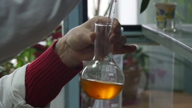 Closeup of a round glass flask kept by a man in a modern biotech laboratory. He pours a yellow fluid into a round flask