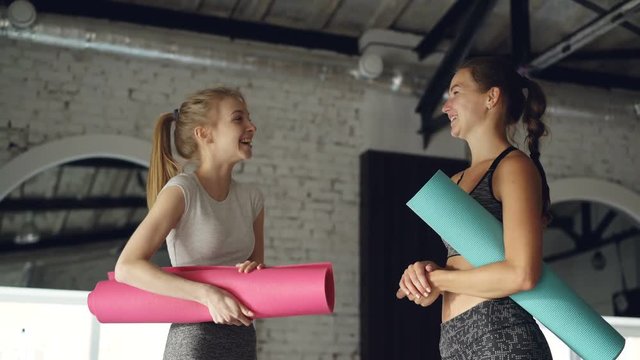 Attractive girls are talking and laughing in large modern sports center. Women are holding colorful yoga mats and wearing fashionable sports clothing.