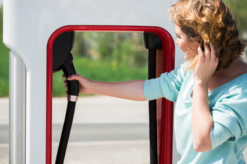 A woman stands at the supercharging station and holds a plug of the charger for an electric car