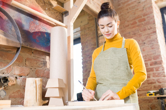Waist up portrait of pretty young woman wearing apron working with wood in crafting shop, standing against brick wall, copy space