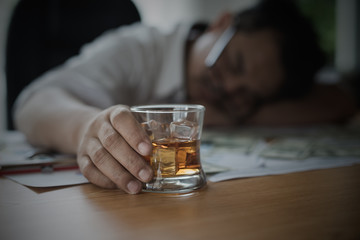 Obraz na płótnie Canvas Stressed asian businessman holding a glass of whiskey. he sleep on the money, data charts, business document at office desk. alcohol addiction - drunk businessman concept
