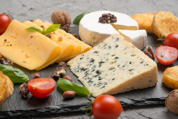 Different kinds of cheeses on a black board, with cherry tomatoes and basil and nuts.