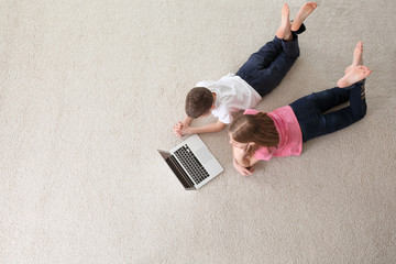 Teenage girl and her brother with laptop lying on cozy carpet at home