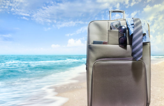 Photo of a travel suitcase packed with shirt, tie, money and sunglasses standing on a sunny beach background.