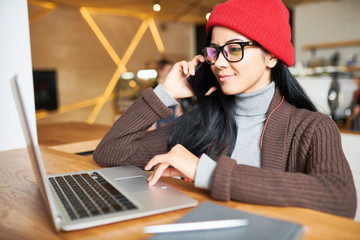 Portrait of trendy young woman speaking by phone and using laptopwhile working  in modern co-working space or cafe, copy space