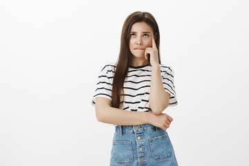 Troubled to made up excuse not to go to university. Confused worried young woman in striped t-shirt, biting lip, holding finger on temple and looking up, thinking, recalling information she forgot