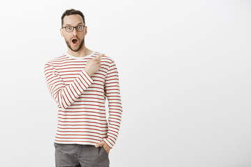 Indoor portrait of pleased impressed handsome man in eyewear, pointing at upper right corner, dropping jaw from positive emotions, asking about product, standing amazed over gray wall