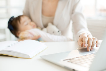 Hand of business mother on laptop keypad during network in office with sleeping baby in the other...