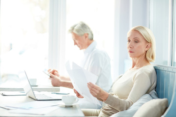 Busy mature woman reading one of financial documents during working day with colleague on background