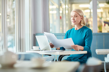 Mature blond woman reading financial documents while sitting by table in cafe and preparing for appointment with client